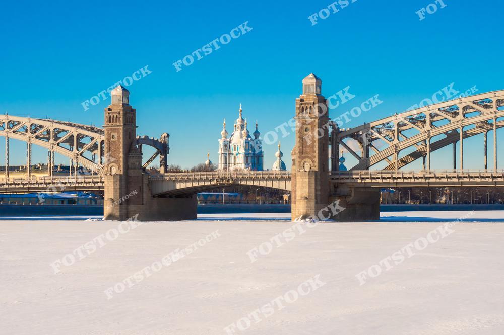 Фотография показывает Большеохтинский мост, соединяющий берега Невы, с видом на величественный Смольный собор. Зимний пейзаж добавляет этому изображению атмосферы спокойствия и красоты северного города.